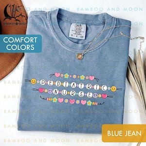May include: A blue jean Comfort Colors t-shirt featuring the text "PEDIATRIC NURSE" in a colourful beaded design. The shirt has a crew neck and short sleeves. A gold necklace with a pendant is draped on the shirt.