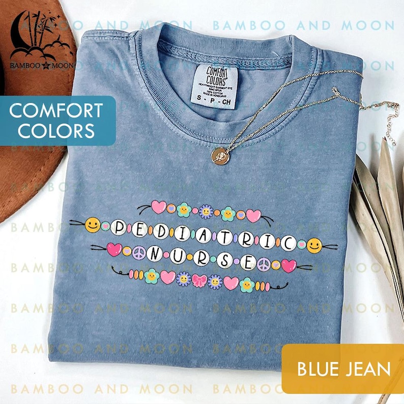 May include: A blue jean Comfort Colors t-shirt featuring the text "PEDIATRIC NURSE" in a colourful beaded design. The shirt has a crew neck and short sleeves. A gold necklace with a pendant is draped on the shirt.