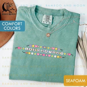 May include: Seafoam green t-shirt with the word "LABORATORY" spelled out in colorful letters and decorative elements. The shirt has a crew neck and short sleeves. A silver necklace with a pendant is draped across the shirt.