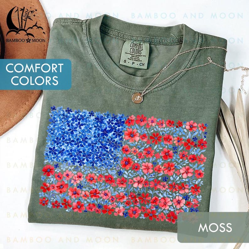 May include: Moss green Comfort Colors t-shirt featuring a floral American flag design. The flag is composed of blue and red flowers. A silver necklace with a pendant is draped on the shirt.