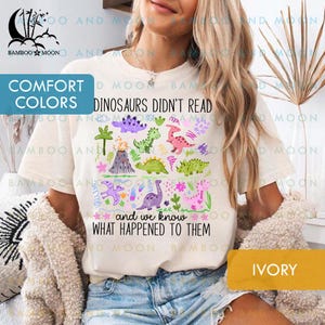 May include: Ivory-colored t-shirt featuring a colorful dinosaur graphic with the text "DINOSAURS DIDN'T READ and we know WHAT HAPPENED TO THEM." The design includes various dinosaur illustrations and floral elements.