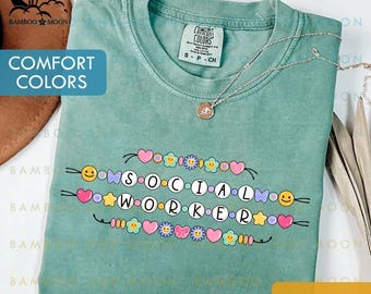 May include: Seafoam green t-shirt with the words "SOCIAL WORKER" in a colorful, beaded design. The shirt features a crew neck and short sleeves. A silver necklace with a pendant rests on the shirt. The shirt is labeled "Comfort Colors".