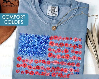 以下が含まれることがあります： 青いジーンズのComfort Colors Tシャツ。花柄のアメリカ国旗のデザイン。旗は青と赤の花で構成されています。ペンダント付きのゴールドネックレスがシャツの上にあります。