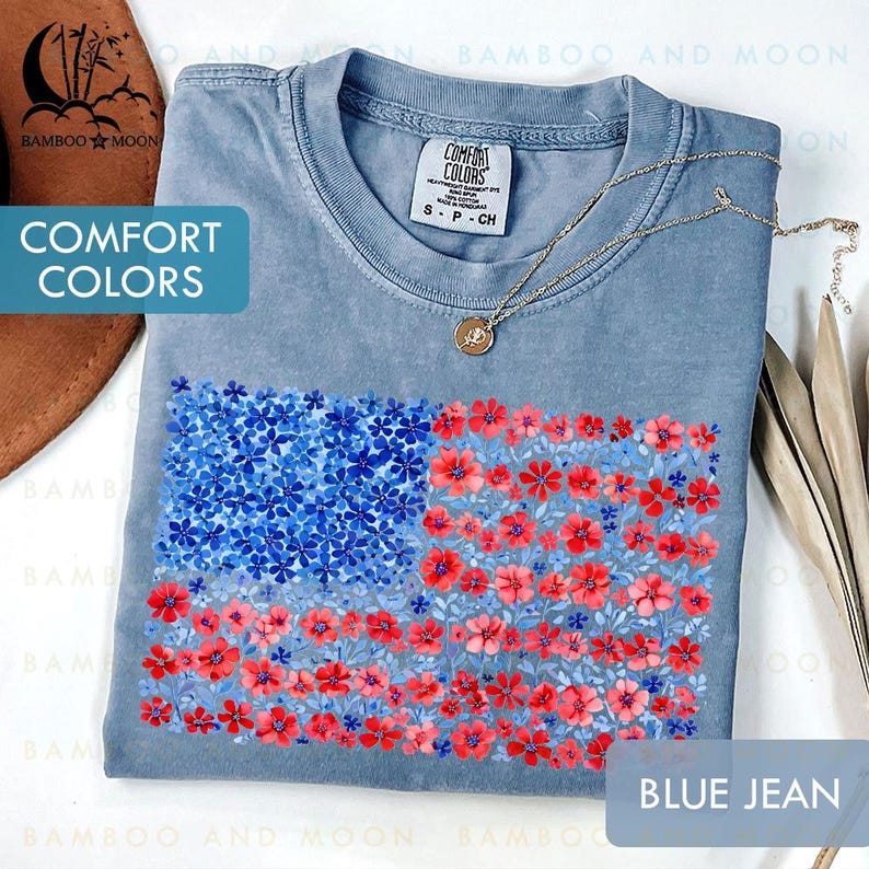 May include: A blue jean Comfort Colors t-shirt featuring a floral American flag design. The flag is composed of blue and red flowers. A gold necklace with a pendant rests on the shirt.