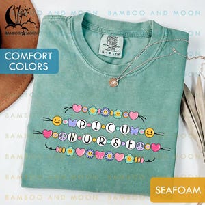 Op de afbeelding: Zeegroen t-shirt met de witte woorden "PICU NURSE", versierd met harten, bloemen en smiley's. Het shirt heeft een ronde hals en korte mouwen. Het merk "Comfort Colors" is zichtbaar.