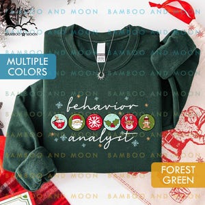 May include: Forest green sweatshirt with the text "behavior analyst" and Christmas-themed graphics. The sweatshirt has a crew neck and long sleeves. The text "Multiple Colors" is also visible.