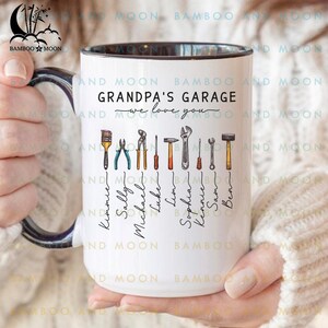 Peut inclure: Mug en céramique blanche avec un bord noir. Il affiche le texte "GRANDPA'S GARAGE" et "we love you", ainsi que des illustrations d'outils et des noms. Le mug est tenu par une personne.