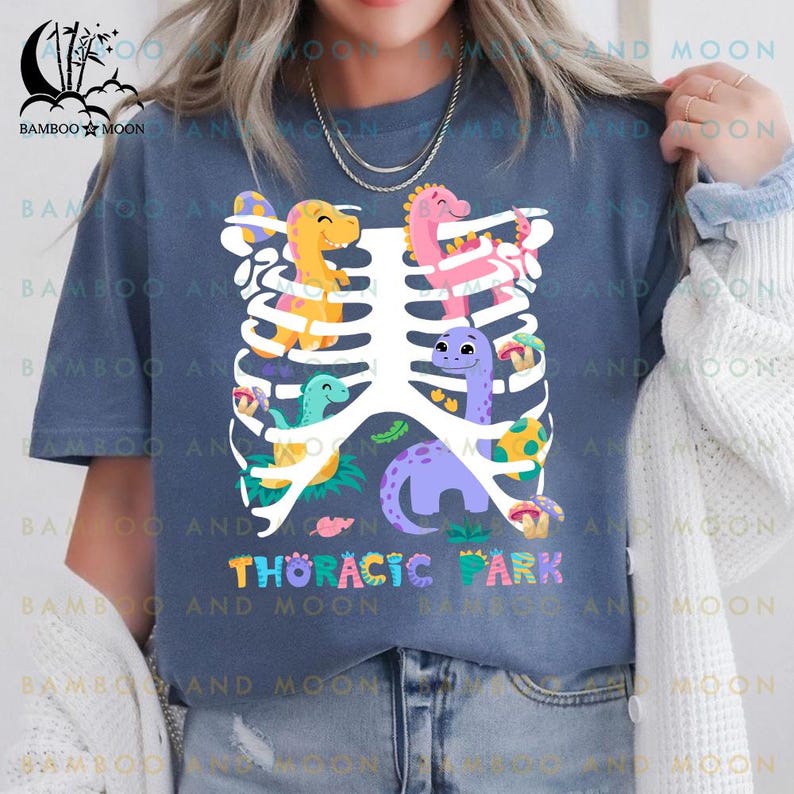 Pu&ograve; includere: Una maglietta blu con una grafica di una gabbia toracica con dinosauri colorati all'interno. Il testo "Thoracic Park" &egrave; stampato sotto la grafica.
