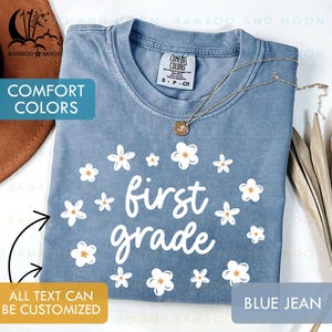May include: A blue jean Comfort Colors t-shirt with the words "first grade" in white script, surrounded by white and orange daisy graphics. A gold necklace with a pendant rests on the shirt. The text "All text can be customized" is also visible.