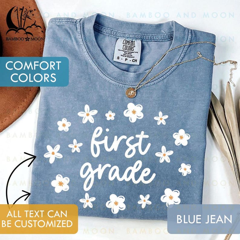May include: A blue jean Comfort Colors t-shirt with the words "first grade" in white script, surrounded by white and orange daisy graphics. A gold necklace with a pendant rests on the shirt. The text "All text can be customized" is also visible.
