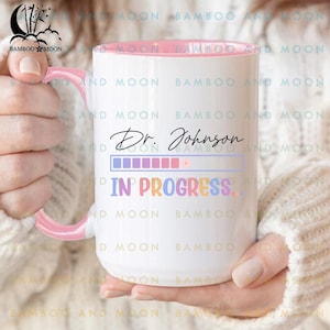 Könnte beinhalten: Weißer Keramikbecher mit rosa Innenraum und Henkel, mit dem Text "Dr. Johnson" über einem Fortschrittsbalken und den Worten "IN PROGRESS" in bunten Buchstaben. Der Becher wird von einer Person gehalten.