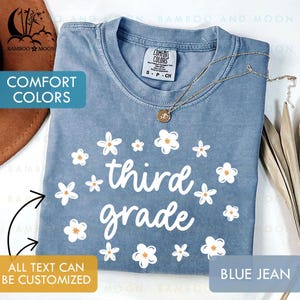 May include: Blue jean Comfort Colors t-shirt with white floral design and the words "third grade" in white. The shirt has a crew neck and short sleeves. A gold necklace with a pendant is on the shirt. The text "ALL TEXT CAN BE CUSTOMIZED" is also visible.