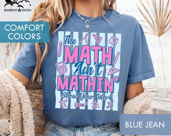 May include: Blue jean colored t-shirt with the text "The MATH Ain't MATHIN' Show Your Work" in pink and white. The shirt features math-related graphics like a calculator, pencil, and ruler. The shirt is a comfort color.
