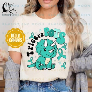 Könnte beinhalten: Ein weißes T-Shirt mit einem türkisfarbenen Triceratops-Motiv und dem Text "Tricera Pots" in türkisfarbener und blauer Schrift. Das Design enthält auch türkisfarbene Bandsymbole.