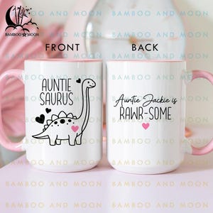 May include: White ceramic mug with a pink handle and rim. The front of the mug features a black dinosaur illustration with the text "AUNTIE SAURUS". The back of the mug says "Auntie Jackie is RAWR-SOME".