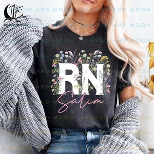May include: A dark gray t-shirt with a white floral design surrounding the letters "RN" and the name "Salim" in pink lettering.