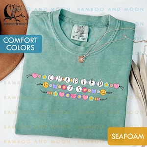May include: Seafoam green Comfort Colors t-shirt with the text "CHAPTER 7.5" in a decorative font, adorned with hearts, flowers, and smiley faces. A silver necklace with a pendant rests on the shirt. The shirt is a size S-CH.