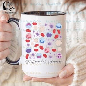 Può includere: Tazza in ceramica bianca con interno e manico neri, con un'illustrazione colorata di cellule del sangue e la scritta "Differentiate Yourself". La tazza è tenuta da una persona.