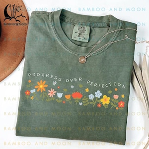 May include: A green t-shirt with a floral design and the text "Progress Over Perfection".