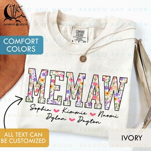 Könnte beinhalten: Elfenbeinfarbenes T-Shirt mit dem Wort "MEMAW" in großen, blütengefüllten Buchstaben. Darunter sind Namen mit Herzen aufgeführt. Das Shirt trägt den Text "COMFORT COLORS" und "ALL TEXT CAN BE CUSTOMIZED". Eine goldene Halskette ist über dem Shirt drapiert.