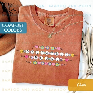 May include: A terracotta-coloured t-shirt featuring the text "PEDIATRIC NURSE" in a beaded design with hearts, flowers, and smiley faces. The shirt has a crew neck and a small Comfort Colors label. A gold necklace is draped on the shirt.