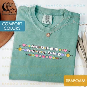 Könnte beinhalten: Meergrünes T-Shirt mit den Worten "SPEECH THERAPY" in einem farbenfrohen, skurrilen Design. Das Shirt hat einen Rundhalsausschnitt und kurze Ärmel. Eine silberne Halskette mit Anhänger ist auf dem Shirt drapiert.