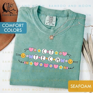 Könnte beinhalten: Meergrünes T-Shirt mit dem Text "COT TECH" in einem farbenfrohen Perlendesign. Das Shirt ist von Comfort Colors und hat eine kleine, runde Anhängerkette. Das Shirt ist in Größe S.