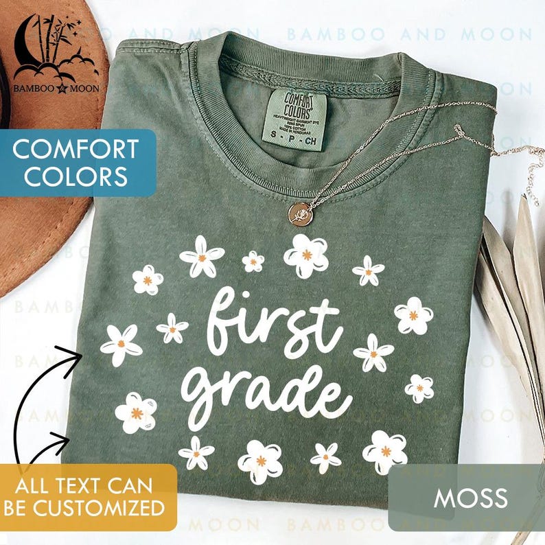 May include: Moss green t-shirt with white floral design and the words "first grade". The shirt has a crew neck and short sleeves. The text "COMFORT COLORS" is visible on the shirt's tag. A gold necklace is draped on the shirt.