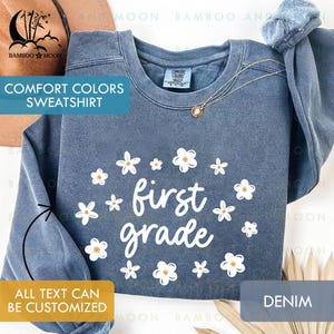 May include: A denim blue Comfort Colors sweatshirt with white floral accents and the words "first grade" in a playful font. The sweatshirt has a crew neck and long sleeves. The text "ALL TEXT CAN BE CUSTOMIZED" is also visible.