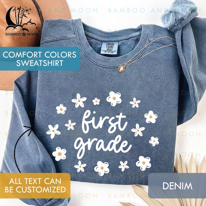 May include: A denim blue Comfort Colors sweatshirt with white floral accents and the words "first grade" in a playful font. The sweatshirt has a crew neck and long sleeves. The text "ALL TEXT CAN BE CUSTOMIZED" is also visible.