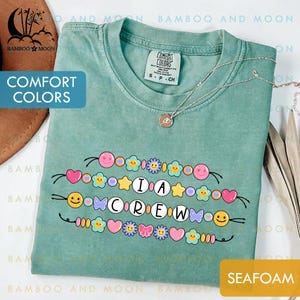 Könnte beinhalten: Meergrünes T-Shirt mit dem Text "I A CREW" in einem verspielten Design mit bunten Smiley-Gesichtern, Herzen, Sternen und Blumen. Das Shirt hat ein Comfort Colors-Etikett und eine zarte Halskette. Das Shirt ist in der Größe S.