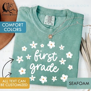 May include: Seafoam green t-shirt with the words "first grade" in white script, surrounded by white flowers with orange centers. The shirt has a Comfort Colors tag and a silver necklace. The text "All text can be customized" is also visible.