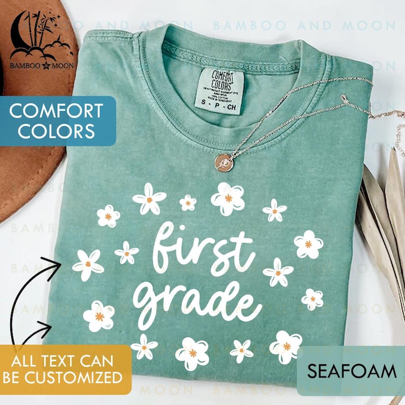 May include: Seafoam green t-shirt with the words "first grade" in white script, surrounded by white flowers with orange centers. The shirt has a Comfort Colors tag and a silver necklace. The text "All text can be customized" is also visible.