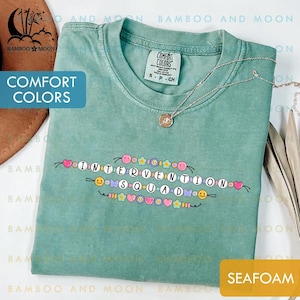 Può includere: T-shirt verde acqua con la scritta "INTERVENTION SQUAD" in un design di perline. La maglietta ha un girocollo e maniche corte. Una collana d'argento con un ciondolo è drappeggiata sulla maglietta. La maglietta è etichettata "Comfort Colors".