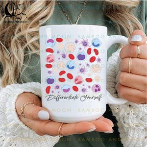 May include: White ceramic mug featuring a colorful illustration of blood cells and the phrase "Differentiate Yourself." The design includes red, purple, blue, and beige cells. The mug has a curved handle and is held by a person.