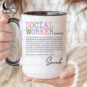 Könnte beinhalten: Weißer Keramikbecher mit schwarzem Rand und Henkel, mit dem Text "SOCIAL WORKER (Substantiv)" in bunten Buchstaben. Der Becher enthält eine Definition eines Sozialarbeiters und den Namen "Sarah" in Schrift.