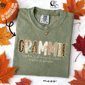 May include: A sage green short-sleeve t-shirt featuring the word GRAMMIE in a patterned font with various autumn-themed designs. Below the word, the names Sophia, Kimmie, Naomi, Dylan, and Dayton are printed in a cursive script with heart icons.