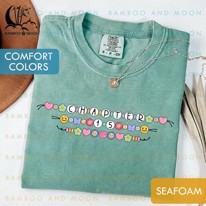 May include: Seafoam green Comfort Colors t-shirt with the text "CHAPTER 15" in a decorative design with hearts, flowers, and smiley faces. A silver necklace with a pendant rests on the shirt.
