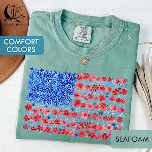 May include: Seafoam green t-shirt with a floral American flag design. The flag features blue flowers in the upper left corner and red flowers in the stripes. The shirt has the words "Comfort Colors" on the tag.