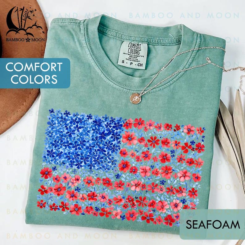 May include: Seafoam green t-shirt with a floral American flag design. The flag features blue flowers in the upper left corner and red flowers in the stripes. The shirt has the words "Comfort Colors" on the tag.