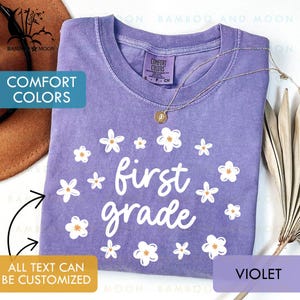 May include: A violet Comfort Colors t-shirt with the words "first grade" in white, surrounded by white and orange-centered flower graphics. A gold necklace with a pendant is draped on the shirt. The text "ALL TEXT CAN BE CUSTOMIZED" is also visible.