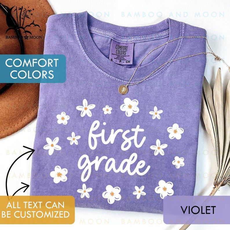 May include: A violet Comfort Colors t-shirt with the words "first grade" in white, surrounded by white and orange-centered flower graphics. A gold necklace with a pendant is draped on the shirt. The text "ALL TEXT CAN BE CUSTOMIZED" is also visible.