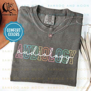 May include: A gray t-shirt with the word "AUDIOLOGY" written in white with a rainbow outline. The shirt is on a table with a wooden surface.