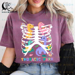 Pu&ograve; includere: Una maglietta viola con un design a gabbia toracica bianca e dinosauri colorati all'interno. Il testo "Thoracic Park" &egrave; stampato sulla maglietta.