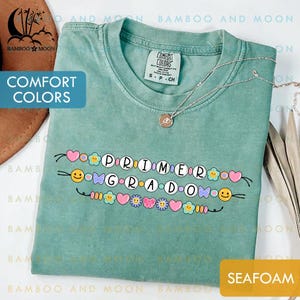 May include: Seafoam green Comfort Colors t-shirt with the words "PRIMER GRADO" in white letters, decorated with colorful hearts, flowers, and smiley faces. A silver necklace with a pendant is draped on the shirt.