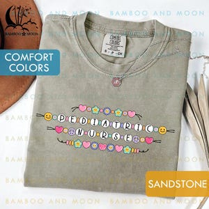 May include: Sandstone-coloured t-shirt featuring the words "PEDIATRIC NURSE" in a colourful design. The shirt has a crew neck and short sleeves. A silver necklace with a small pendant is on the shirt.