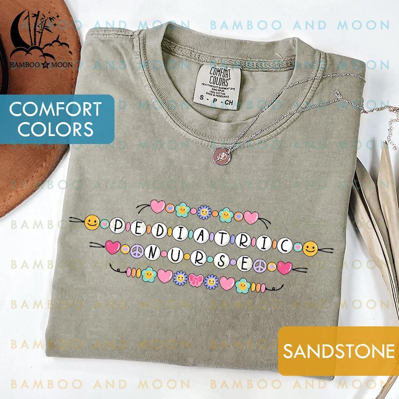 May include: Sandstone-coloured t-shirt featuring the words "PEDIATRIC NURSE" in a colourful design. The shirt has a crew neck and short sleeves. A silver necklace with a small pendant is on the shirt.