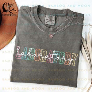 Op de afbeelding: Een donkergrijs T-shirt met het woord "LABORATORY" erop geschreven in regenboogkleuren. Het shirt is gemaakt van een zacht, comfortabel materiaal en heeft een ontspannen pasvorm.