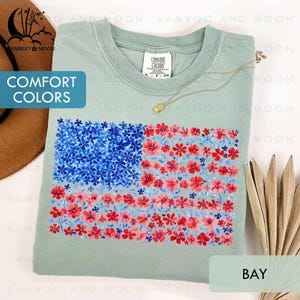 May include: A light green t-shirt with a floral American flag design. The flag features blue flowers in the upper left corner and red and pink flowers for the stripes. The shirt has the text "COMFORT COLORS" and "BAY".