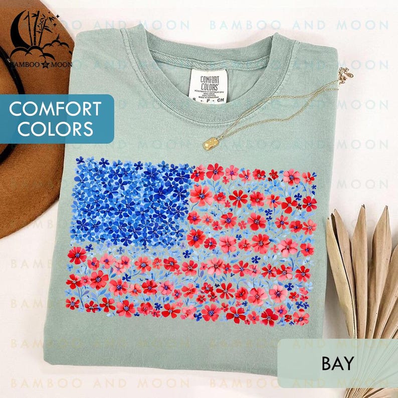 May include: A light green t-shirt with a floral American flag design. The flag features blue flowers in the upper left corner and red and pink flowers for the stripes. The shirt has the text "COMFORT COLORS" and "BAY".